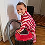 child, toddler, baby, pacifier, vacuum_cleaner, striped_pajamas, red_and_white_stripes, hand_on_object, wooden_floor, door, shadow, smile, curious, home_interior, appliance, standing, portrait, playful, furniture, flooring