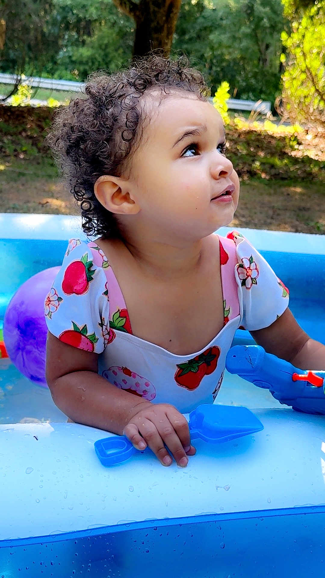 Anastara is registered to the contest to win money with this photo: child, toddler, curly_hair, inflatable_pool, water, water_toy, blue_shovel, ball, swimsuit, strawberry_print, cute, outdoor, summer, portrait, looking_up, playtime, hand, skin, grass, tree
