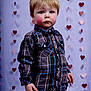 blond_hair, boy, button_up, carpet_floor, casual_clothing, child, cute, expressionless, hands, hanging_hearts, indoor, jeans, plaid_shirt, portrait, rosy_cheeks, small_person, standing, studio_backdrop, toddler, valentine_theme