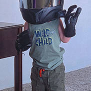 Samuel is registered to the contest to win money with this photo: child, helmet, gloves, tank_top, shorts, sandals, carpet, furniture, indoor, standing, wild_child, playful, oversized, reflection, footwear, clothing, person, home, cute, pretend_play