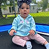 baby, child, girl, headband, bow, blue_hoodie, pink_pants, socks, trampoline, outdoor, fence, grass, curious, portrait, sitting, cute, infant, young_child, daylight, play