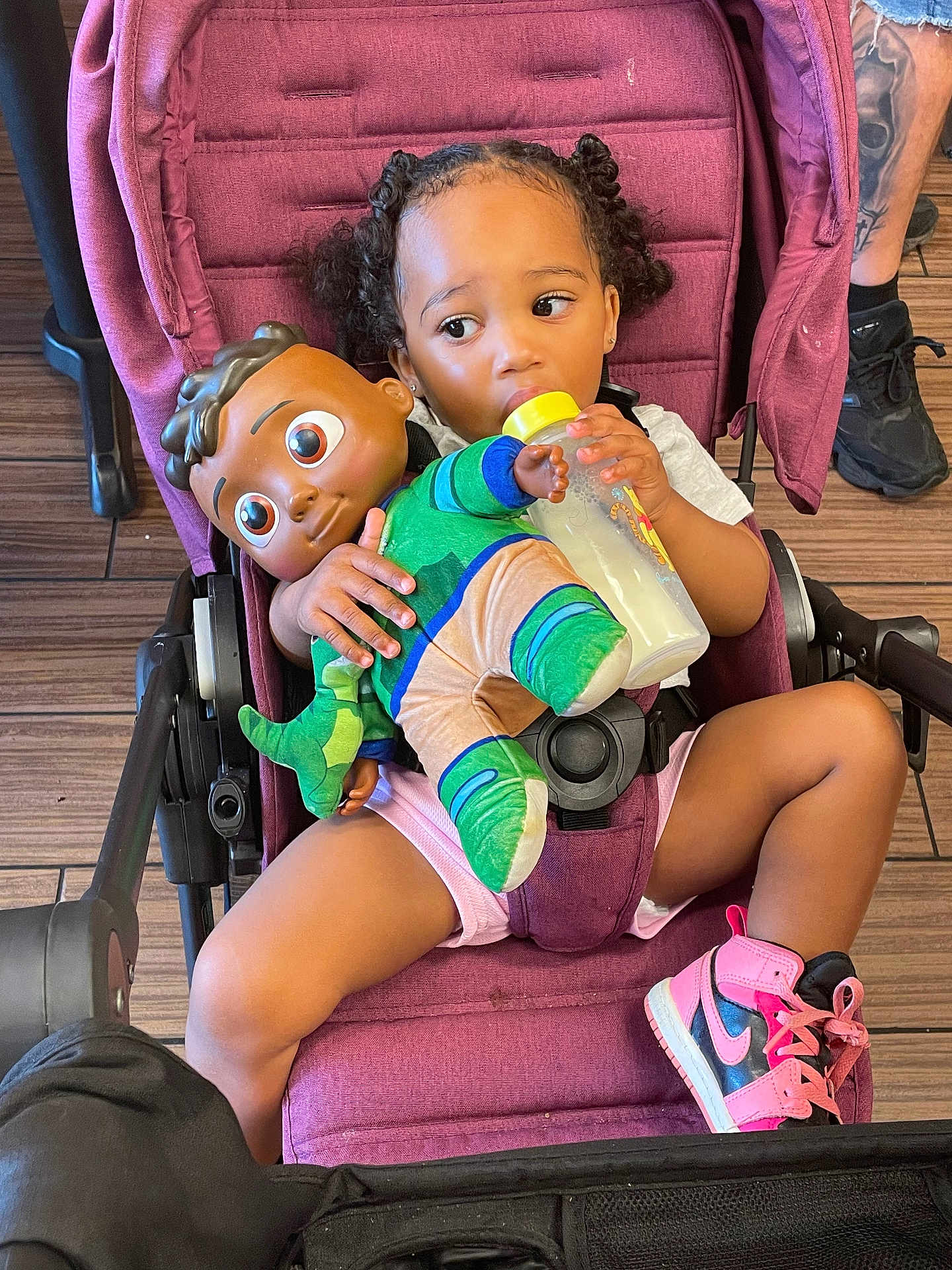 Layla Salley is registered to the contest to win money with this photo: toddler, stroller, baby_bottle, doll, pink_sneakers, child, toy, floor, wooden_floor, person, clothing, hair, seat, footwear, casual, indoor, holding, sitting, cute, playtime