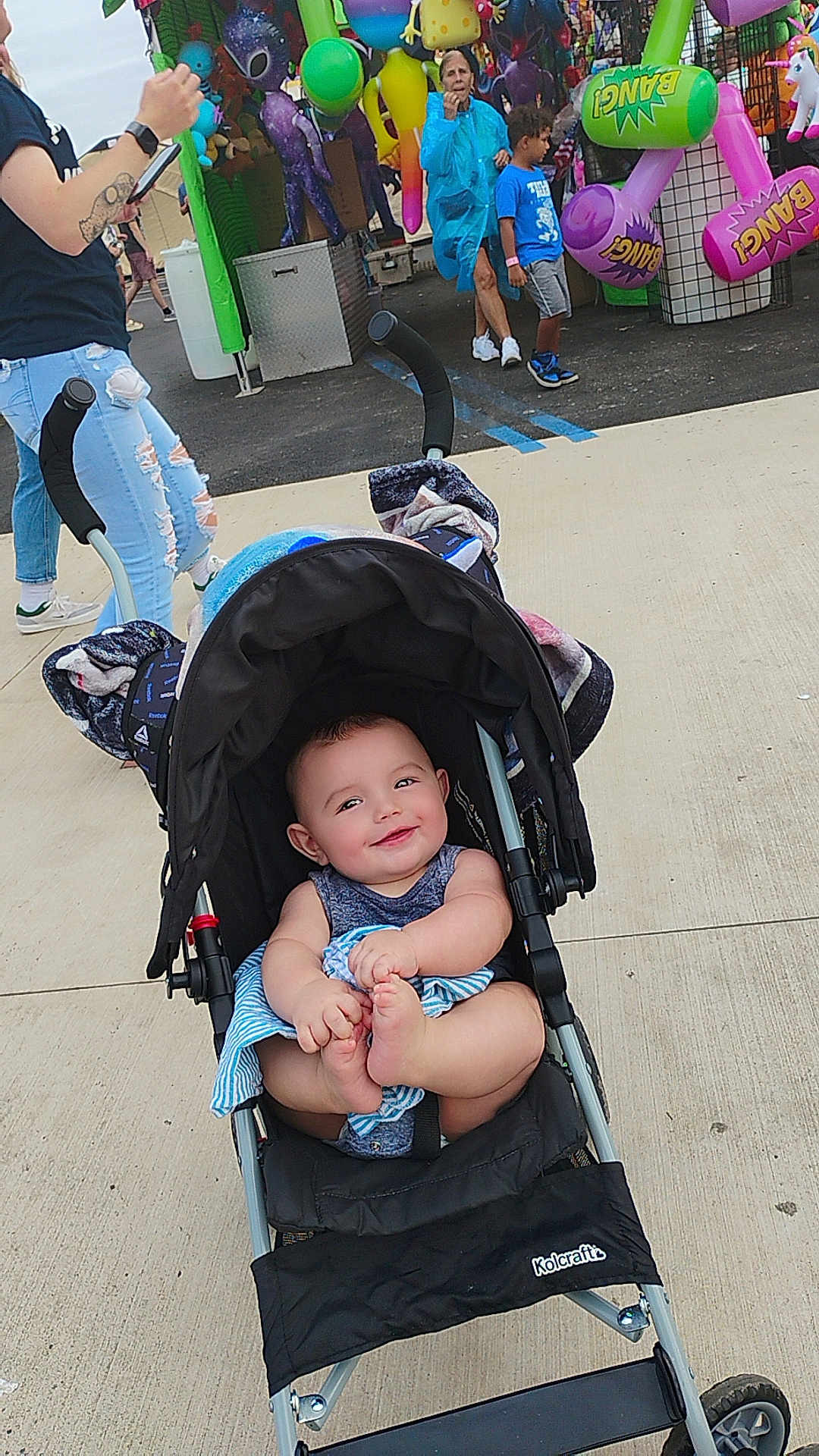 Isaiah is registered to the contest to win money with this photo: baby, stroller, child, smiling, outdoor, carnival, inflatable_toys, people, sidewalk, casual_clothing, happy, face, footwear, hand, toy, event, fun, daytime, urban, crowd