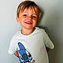child, boy, smiling, tshirt, cartoon, stitch, ice_cream, light_hair, portrait, casual, standing, wall, indoor, happy, young, cute, clothing, expression, person, background