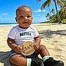 baby, child, tropical_beach, palm_trees, ocean, sand, blue_sky, sitting, smiling, portrait, infant, milestone_sign, wooden_sign, white_shirt, shorts, sneakers, socks, outdoors, vacation, sunlight