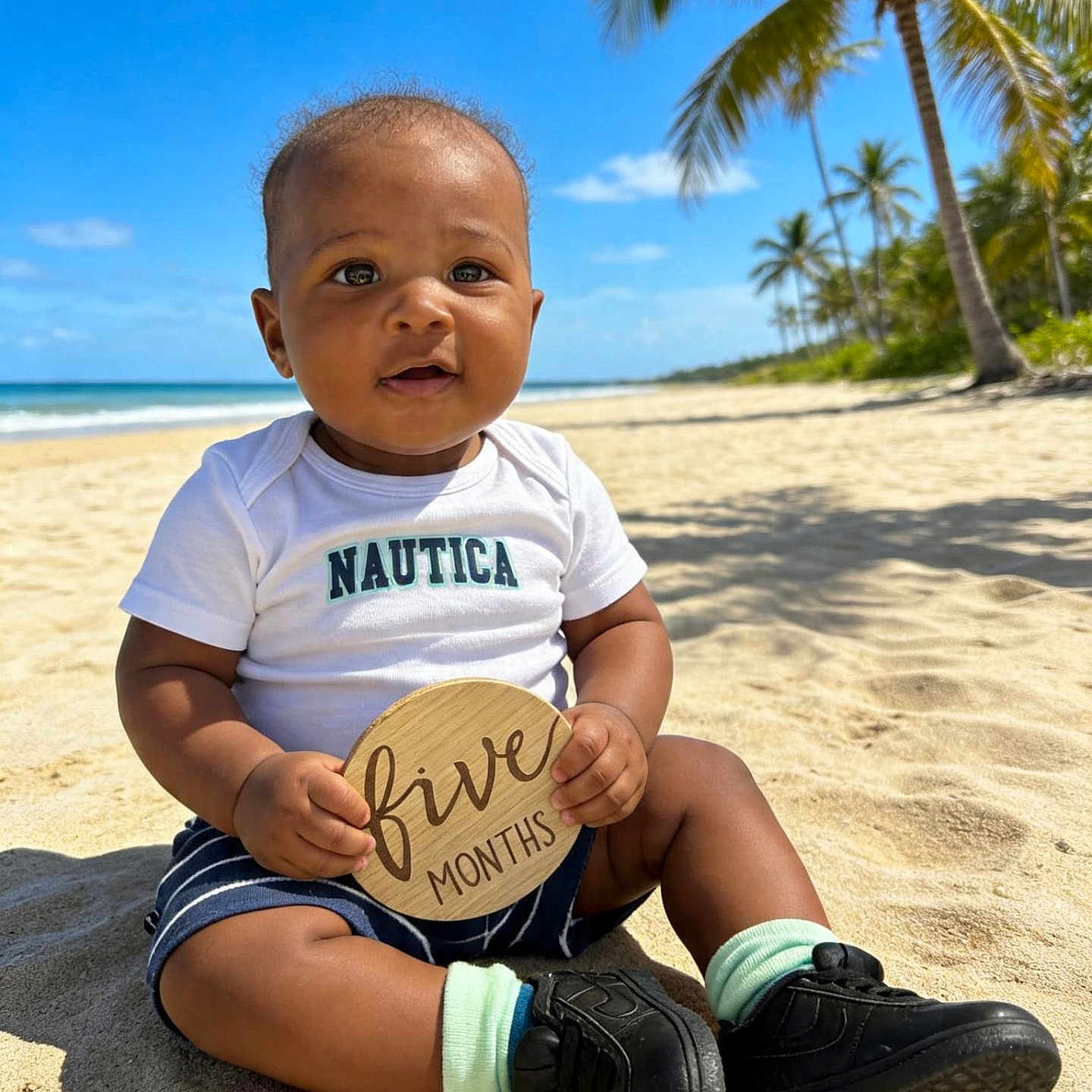 Kaimani joined the competition — help win amazing prizes! baby, child, tropical_beach, palm_trees, ocean, sand, blue_sky, sitting, smiling, portrait, infant, milestone_sign, wooden_sign, white_shirt, shorts, sneakers, socks, outdoors, vacation, sunlight