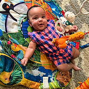 Maela joined the competition — help win amazing prizes! activity_gym, baby, carpet, checkered_clothing, child, colorful, face, happy, indoor, infant, lying_down, nursery, overhead_view, play_mat, playtime, portrait, romper, smiling, stuffed_animal, toys