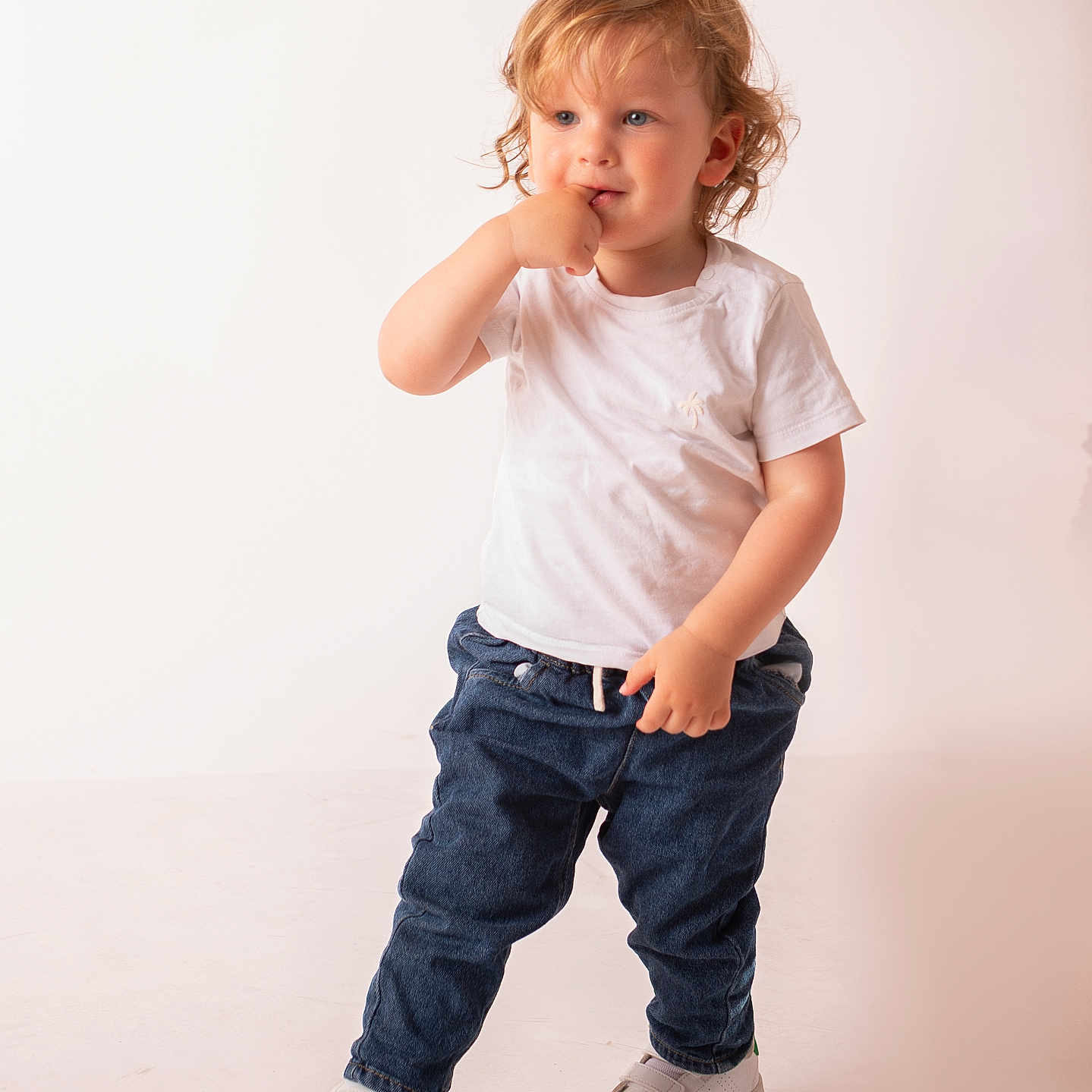 Leo participe au concours pour gagner de l'argent avec cette photo : blue_jeans, casual_clothing, child, curious, curly_hair, cute, fashion, finger_in_mouth, indoor, light_background, one_foot, person, playful, portrait, standing, studio, toddler, white_shoes, white_tshirt, young_child