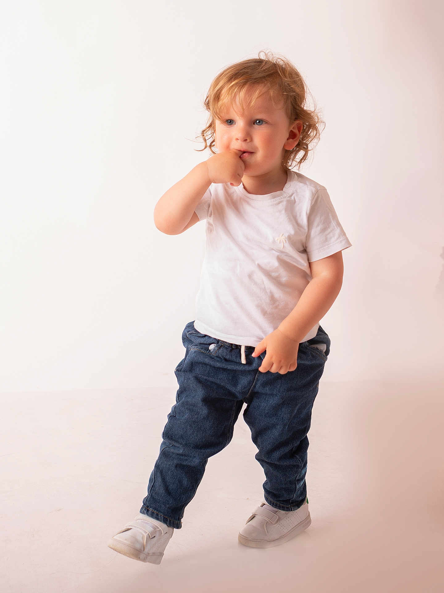 Leo participe au concours pour gagner de l'argent avec cette photo : blue_jeans, casual_clothing, child, curious, curly_hair, cute, fashion, finger_in_mouth, indoor, light_background, one_foot, person, playful, portrait, standing, studio, toddler, white_shoes, white_tshirt, young_child