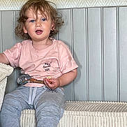 Leo a rejoint le concours — aidez-le/la à gagner de superbes lots ! toddler, child, curly_hair, pink_shirt, grey_pants, sitting, couch, snack, indoor, person, blue_eyes, wall_paneling, casual_clothing, cute, comfortable, young_child, expression, home, relaxed, furniture