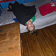 Arian joined the competition — help win amazing prizes! child, bed, blanket, pillow, wooden_floor, table, room, indoor, upside_down, sleepy, wall_art, text, green_shirt, red_pillow, black_blanket, head, face, person, furniture, floor