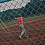 action, athlete, baseball, bat, chain_link_fence, child, dirt, fence, field, grass, helmet, jersey, number, outdoor, recreation, sky, sport_equipment, sports, swing, trees