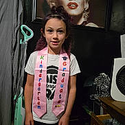 Davina is registered to the contest to win money with this photo: child, kindergarten, graduate, pink_sash, portrait, marilyn_monroe, vacuum_cleaner, indoor, tshirt, standing, hair_tied, drawer, clutter, wooden_floor, face, person, smile, celebration, home, decor