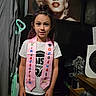 child, kindergarten, graduate, pink_sash, portrait, marilyn_monroe, vacuum_cleaner, indoor, tshirt, standing, hair_tied, drawer, clutter, wooden_floor, face, person, smile, celebration, home, decor