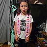 child, kindergarten_graduate, pink_sash, white_tshirt, black_pants, vacuum_cleaner, marilyn_monroe_poster, indoor, wooden_floor, dresser, curtain, celebration, smiling, portrait, person, hair_tied_up, home_interior, floor, achievement, young
