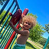 child, toddler, curly_hair, playground, outdoor, sunny, blue_sky, red_panel, green_shorts, summer, park, metal_bars, happy, play, nature, grass, tree, daytime, smile, person
