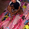 child, boy, smiling, sitting, blanket, barbie, toy, christmas_tree, decorations, lights, floor, indoors, casual_clothing, shorts, tshirt, playful, holiday, festive, present, happy