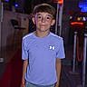 child, boy, purple_shirt, shorts, red_carpet, night, neon_lights, indoor, casual_clothing, smiling, standing, person, portrait, urban, fashion, youth, confident, leisure, event, background