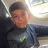 child, boy, airplane, window, seat, sunlight, smile, clothing, jacket, nike_logo, airport, travel, young, casual, portrait, indoors, seatbelt, technology, device, relaxed