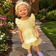Ocean Rae is registered to the contest to win money with this photo: child, toddler, yellow_dress, headband, smiling, outdoor, garden, flowers, greenery, stone_pathway, wooden_gate, sunlight, happy, cute, curly_hair, purple_shoes, nature, summer, portrait, person