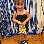 Alessia is registered to the contest to win money with this photo: toddler, child, snack, black_dress, pigtails, indoor, wooden_floor, party_decor, streamers, shoes, black_and_white_shoes, standing, happy, cute, young_child, person, celebration, fun, background, hair