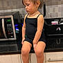 toddler, child, kitchen, counter, black_outfit, sneakers, ponytail, pout, serious, indoor, cabinet, microwave, tile_wall, sitting, white_socks, modern_kitchen, young_child, casual, portrait, cute