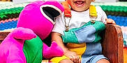 Jorel joined the competition — help win amazing prizes! toddler, child, smiling, plush_toy, barney, bench, colorful_clothing, overalls, white_shoes, indoor, carpet, television, animation, happy, cute, playful, toy, furniture, portrait, baby