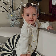 Layan is registered to the contest to win money with this photo: baby, child, toddler, standing, smiling, portrait, indoors, sofa, cushion, cream_clothing, yellow_socks, hair_bows, floral_decoration, decorative_tree, living_room, furniture, home, patterned_pillow, happy, photography