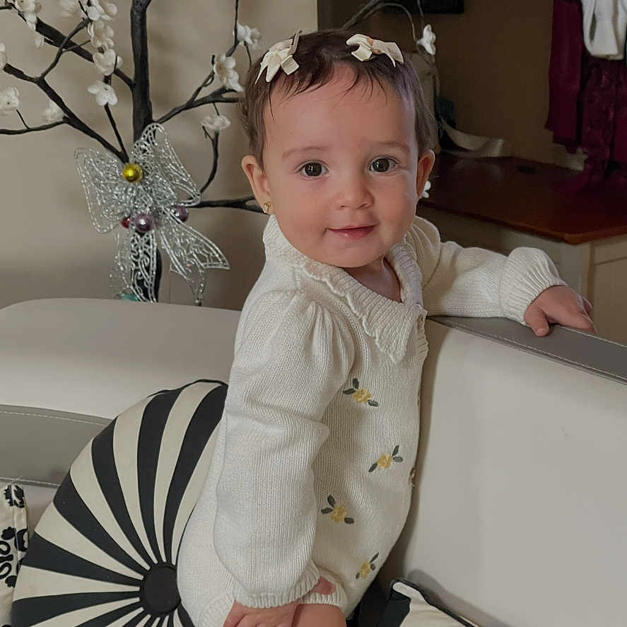 Layan is registered to the contest to win money with this photo: baby, child, toddler, standing, smiling, portrait, indoors, sofa, cushion, cream_clothing, yellow_socks, hair_bows, floral_decoration, decorative_tree, living_room, furniture, home, patterned_pillow, happy, photography