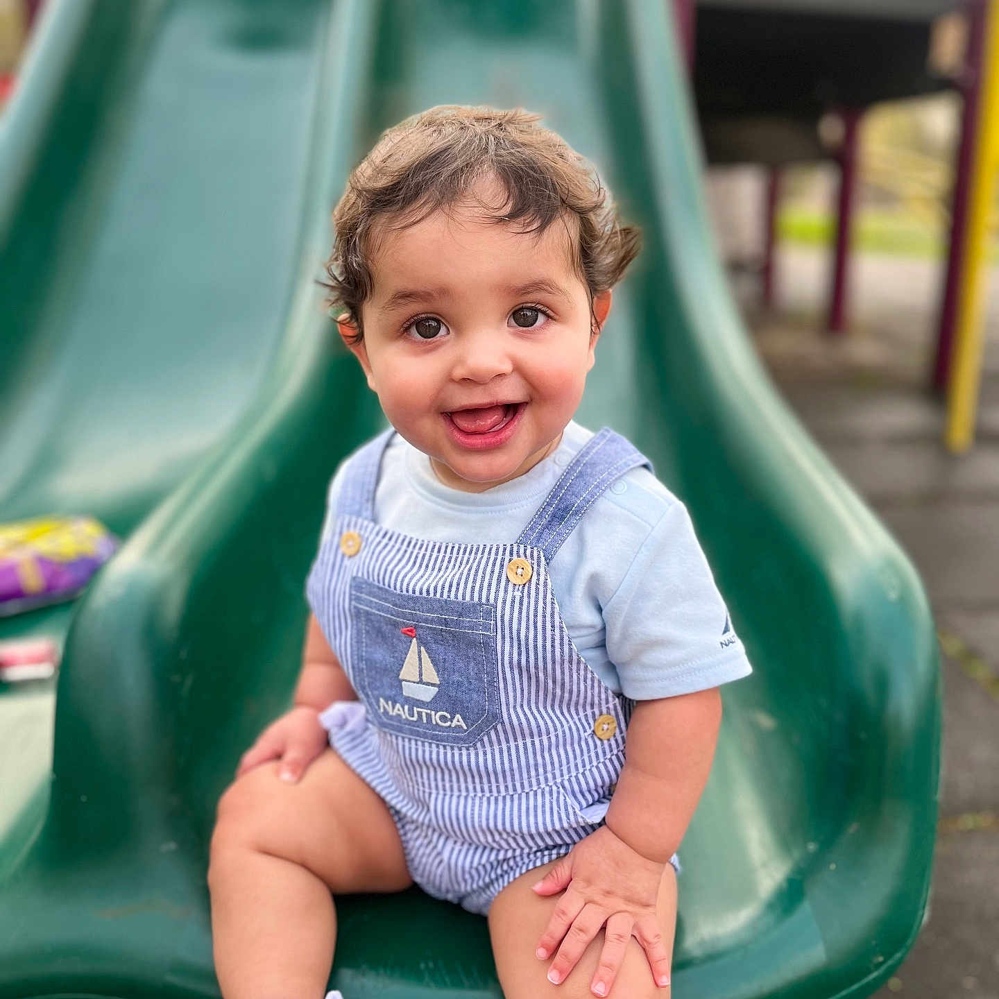 Jordel is registered to the contest to win money with this photo: baby, clothing, face, footwear, grass, happy, head, outdoorplayarea, outdoors, pants, person, photography, plant, playarea, portrait, shoe, sitting, slide, smile, toy