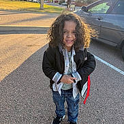 Brodie joined the competition — help win amazing prizes! child, curly_hair, smiling, jacket, jeans, sneakers, parking_lot, car, sunlight, outdoor, person, casual_clothing, street, daytime, young_child, happy, standing, fashion, urban, nature