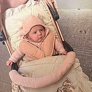 Blossom is registered to the contest to win money with this photo: baby, infant, stroller, pink, knit_hat, knit_sweater, face, outdoor, child, soft_fabric, bow, blanket, padded, cozy, person, cute, small, seat, warm_clothing, curious