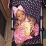 baby, child, stroller, pink_dress, headband, shoe, polka_dots, indoor, floor, curious, seated, footwear, person, toddler, fabric, pattern, toy, hand, face, infant