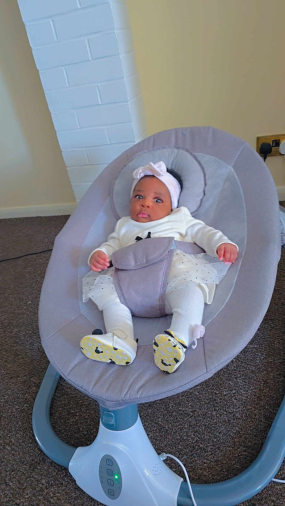 Lael is registered to the contest to win money with this photo: baby, infant, child, baby_rocker, headband, white_clothing, carpet, indoor, seat, footwear, baby_shoes, modern, cushion, curious, young_child, portrait, cute, comfortable, floor, plug_socket