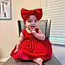 baby, child, red_dress, headband, bow, smiling, sitting, indoor, chair, cute, happy, finger_in_mouth, portrait, infant, person, cute_expression, soft_lighting, window, blinds, furniture