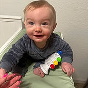 Camryn is registered to the contest to win money with this photo: baby, child, smiling, toy, gray_clothing, indoor, hand, nail_polish, colorful, fingernails, face, cute, infant, reaching, closeup, person, skin, foot, crawling, green