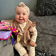 Kehlani joined the competition — help win amazing prizes! baby, smiling, child, pajamas, headband, knitted_vest, plush_toy, colorful_toy, couch, gray_couch, pillow, fluffy_pillow, blanket, patterned_blanket, indoors, happy, seated, person, cute, cozy