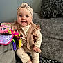 baby, smiling, child, pajamas, headband, knitted_vest, plush_toy, colorful_toy, couch, gray_couch, pillow, fluffy_pillow, blanket, patterned_blanket, indoors, happy, seated, person, cute, cozy