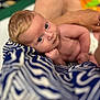 baby, infant, blue_eyes, blanket, patterned_blanket, skin, arm, hand, person, resting, curious, close_up, portrait, indoor, soft_light, head, face, newborn, support, human