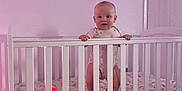 Carson is registered to the contest to win money with this photo: baby, crib, infant, bedroom, hand, mattress, pajamas, white_crib, standing, baby_toys, soft_lighting, indoors, child, person, sleep_area, furniture, pink_light, looking, holding, home