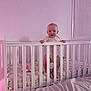 baby, crib, infant, bedroom, hand, mattress, pajamas, white_crib, standing, baby_toys, soft_lighting, indoors, child, person, sleep_area, furniture, pink_light, looking, holding, home