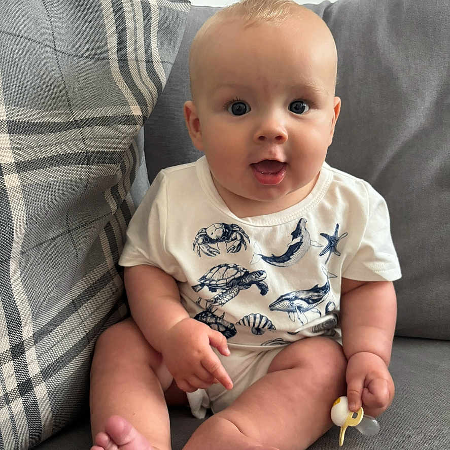 Carson is registered to the contest to win money with this photo: baby, child, chubby, couch, curious, cute, face, feet, hands, homedecor, indoor, infant, marine_animals, onesie, pacifier, plaid_pillow, sitting, smiling, soft_fabric, white_clothing