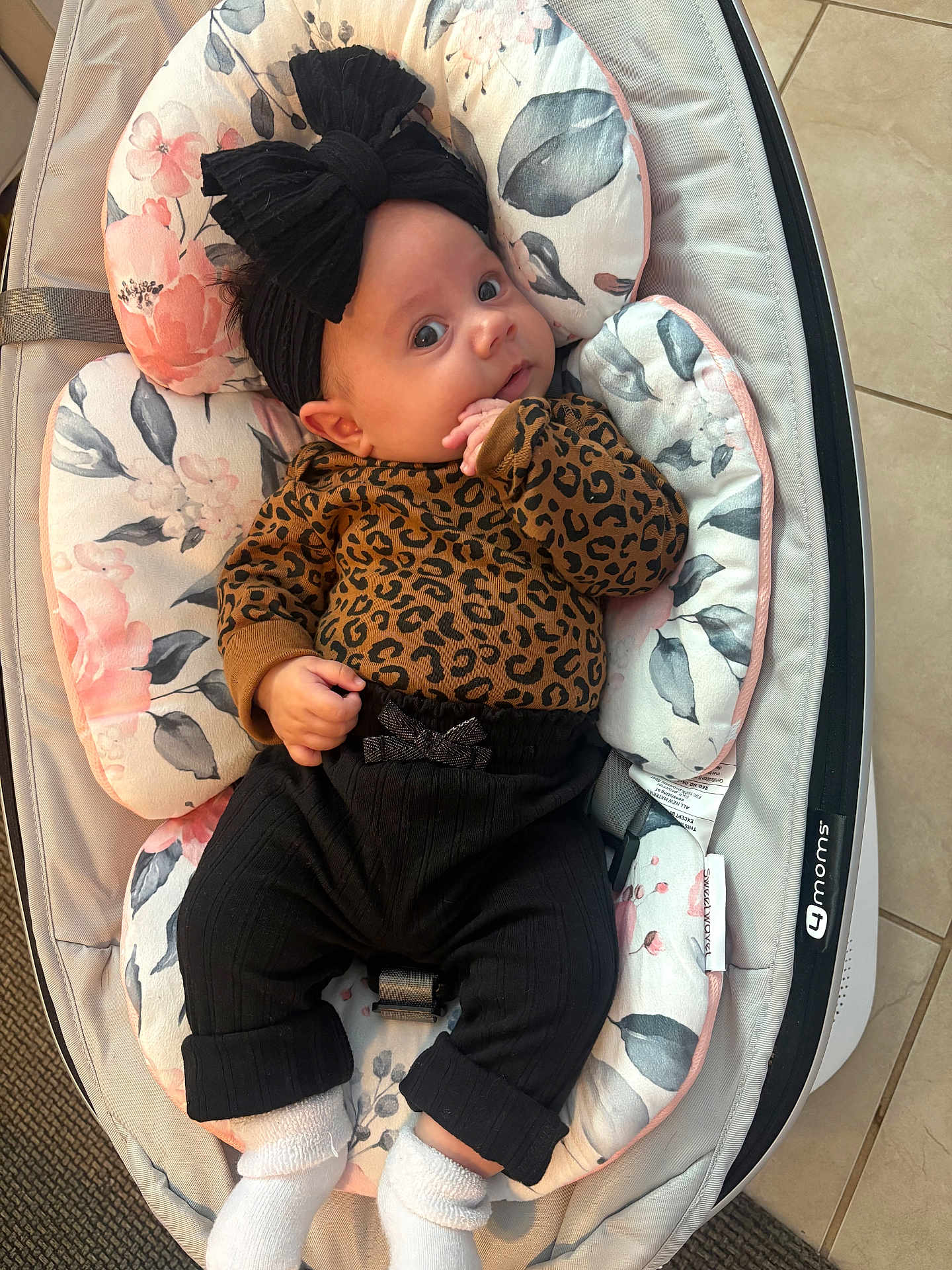 Alana is registered to the contest to win money with this photo: baby, infant, child, black_bow, leopard_print, pants, socks, baby_seat, floral_pattern, cushion, indoor, floor_tiles, cute, portrait, person, young_child, comfort, resting, clothing, headwear