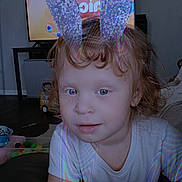 Zemyrah is registered to the contest to win money with this photo: child, bunny_ears, curly_hair, indoor, television, toy, blanket, white_shirt, face, person, living_room, floor, furniture, couch, hair, smile, playful, cute, home, portrait