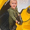 Kolt is registered to the contest to win money with this photo: toddler, child, smile, yellow_car, ride_on_toy, hoodie, green_clothing, indoor, mall, tile_floor, short_hair, footwear, cute, playful, vehicle, hands, standing, young_child, boy, curious