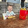 Kolt is registered to the contest to win money with this photo: child, toddler, smiling, table, fast_food, fries, chicken_nuggets, drink, happy, living_room, couch, lamp, wall_art, clock, red_box, mcdonalds, wooden_table, indoor, person, home