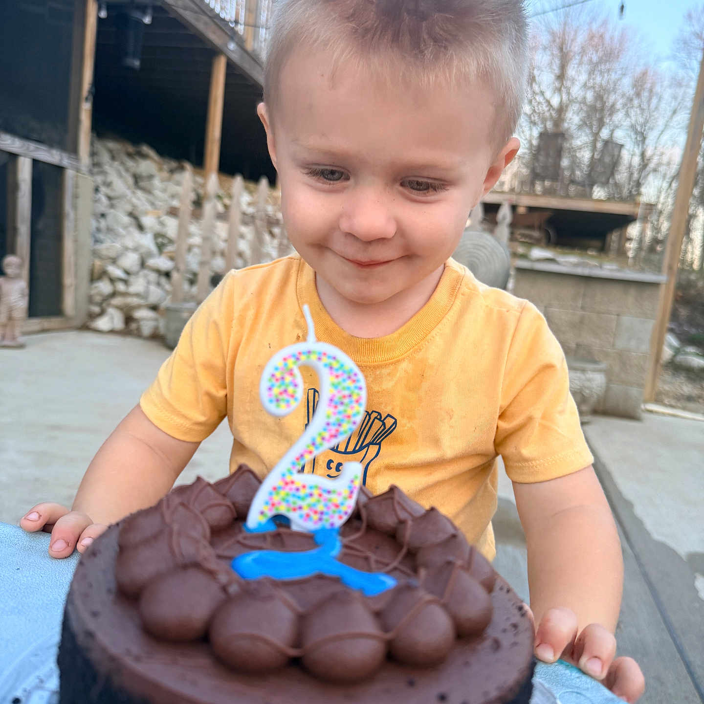 Kolt is registered to the contest to win money with this photo: child, toddler, birthday_cake, chocolate_cake, candle, number_two, yellow_shirt, outdoor, celebration, smiling, happy, face, short_hair, table, concrete, wooden_deck, rocks, daylight, nature, background