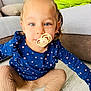 toddler, child, pacifier, blue_eyes, blue_top, floral_pattern, beige_tights, sitting, wooden_floor, living_room, couch, blanket, indoor, cute, portrait, young_child, smiling, person, home, cozy