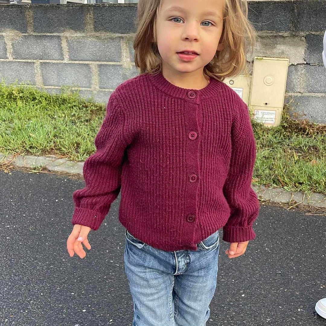 Chloé participe au concours pour gagner de l'argent avec cette photo : blonde_hair, blue_jeans, casual_clothing, child, curious_expression, daylight, fence, footwear, grass, house, maroon_sweater, outdoor, paved_road, person, portrait, standing, stone_wall, white_sneakers, windows, young_child