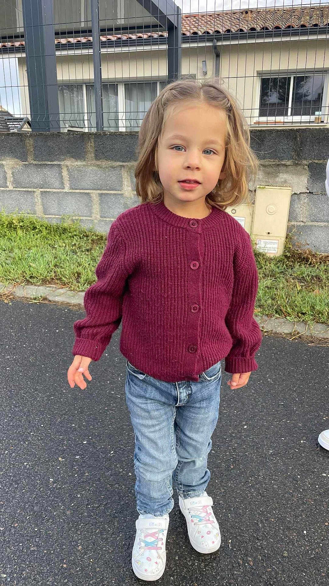 Chloé participe au concours pour gagner de l'argent avec cette photo : child, young_child, blonde_hair, maroon_sweater, blue_jeans, white_sneakers, standing, outdoor, paved_road, grass, stone_wall, house, windows, fence, curious_expression, casual_clothing, daylight, portrait, person, footwear